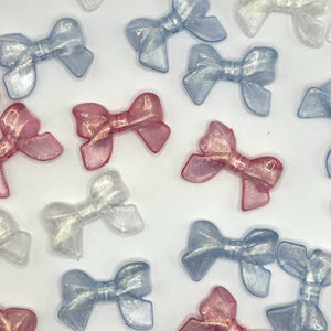 May include: Assortment of small, iridescent bows in blue, pink, and white. The bows are made of a translucent material with a shimmery finish.