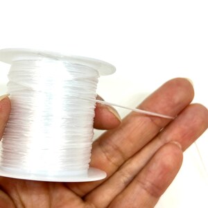 Pu&ograve; includere: Un rocchetto di filo elastico trasparente per la creazione di gioielli. Il filo &egrave; avvolto attorno a un rocchetto di plastica bianca e viene tenuto in una mano.