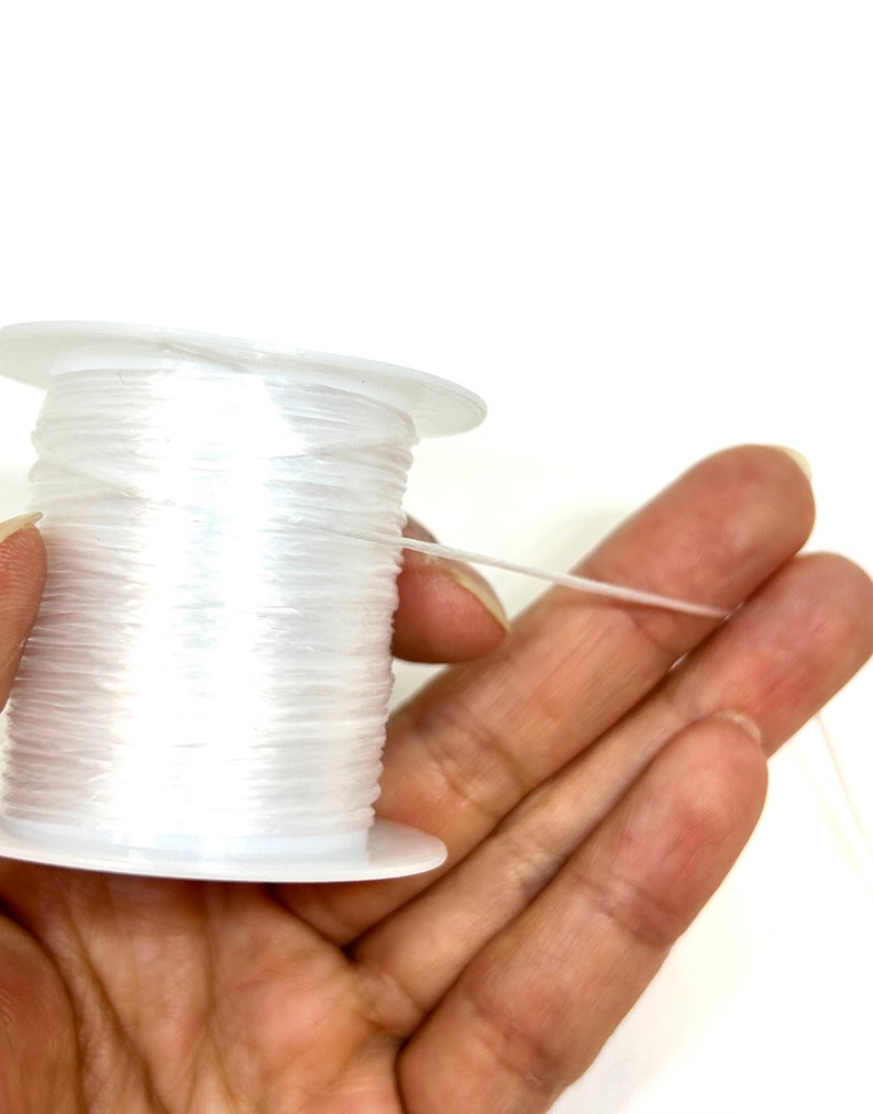 Pu&ograve; includere: Un rocchetto di filo elastico trasparente per la creazione di gioielli. Il filo &egrave; avvolto attorno a un rocchetto di plastica bianca e viene tenuto in una mano.