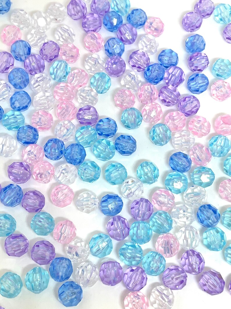 May include: A collection of faceted plastic beads in various shades of blue, pink, and clear. The beads are scattered on a white background.