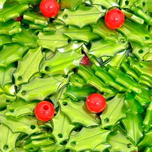 May include: A large pile of green holly leaf charms with red beads scattered throughout. The charms are shiny and have a metallic finish.