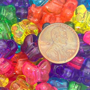 May include: A close-up of a large pile of colorful plastic butterfly-shaped beads. The beads are in various shades of red, orange, yellow, green, blue, purple, and pink. A US penny is visible in the center of the image for scale.