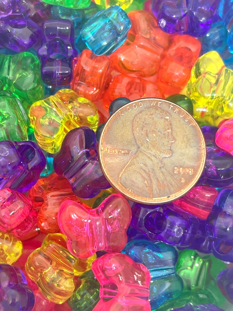 May include: A close-up of a large pile of colorful plastic butterfly-shaped beads. The beads are in various shades of red, orange, yellow, green, blue, purple, and pink. A US penny is visible in the center of the image for scale.