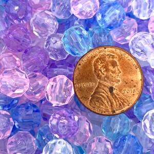 May include: A close-up of a large quantity of faceted plastic beads in shades of purple, blue, and pink. A US penny is included for scale.