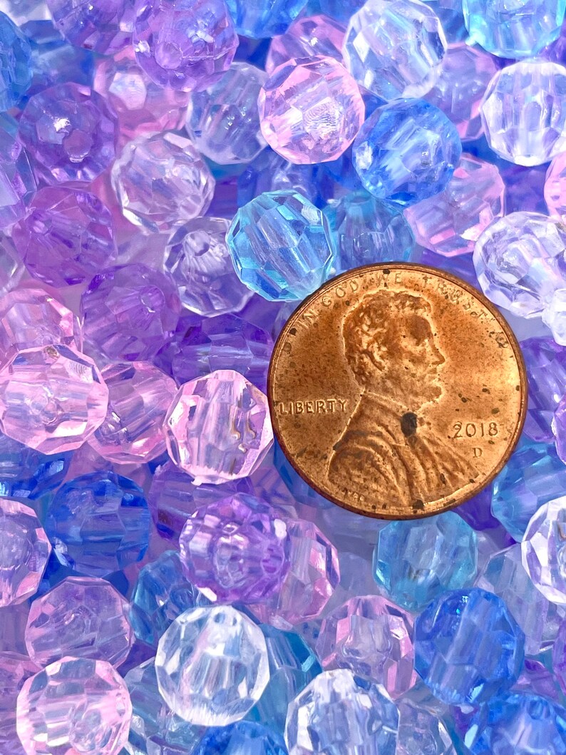 May include: A close-up of a large quantity of faceted plastic beads in shades of purple, blue, and pink. A US penny is included for scale.