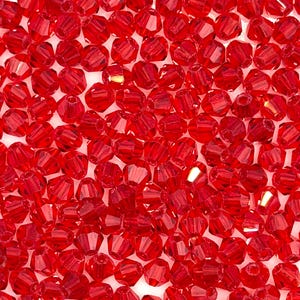May include: A close-up shot of numerous faceted, red beads. The beads are small, with multiple facets that catch the light, creating a sparkling effect. The beads are scattered across a white surface, suggesting they are for jewelry making or crafts.