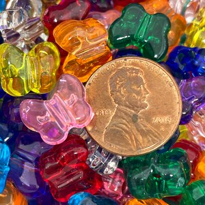 Pu&ograve; includere: Primo piano di una variet&agrave; di perline a forma di farfalla in plastica colorata. Le perline sono di vari colori, tra cui rosso, arancione, giallo, verde, blu, viola e trasparente. Tutte le perline hanno la forma di una farfalla. C'&egrave; una moneta di rame al centro dell'immagine.