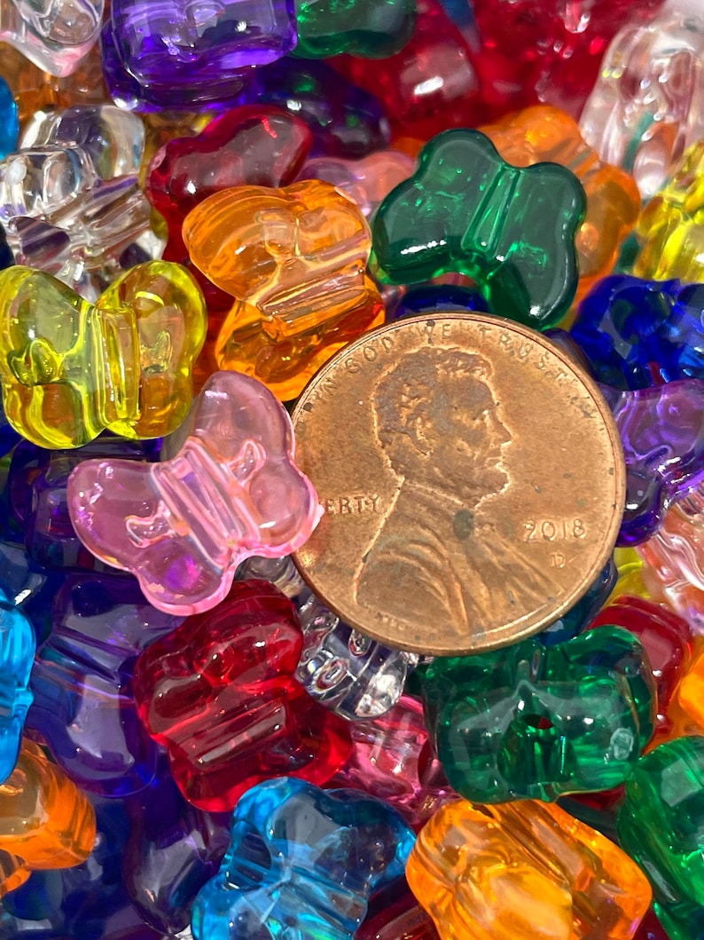Pu&ograve; includere: Primo piano di una variet&agrave; di perline a forma di farfalla in plastica colorata. Le perline sono di vari colori, tra cui rosso, arancione, giallo, verde, blu, viola e trasparente. Tutte le perline hanno la forma di una farfalla. C'&egrave; una moneta di rame al centro dell'immagine.