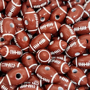 May include: A large group of brown and white football-shaped beads. The beads have a white stitching detail and a hole through the center.