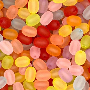 May include: A close-up shot of a collection of colorful, oval-shaped beads. The beads are in various shades of orange, yellow, pink, red, and purple. Each bead has a small hole, suggesting they are for stringing. The beads are scattered, creating a vibrant, textured surface.