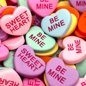 May include: A close-up of a pile of heart-shaped sweets with various messages, including "Be Mine", "Sweet Heart", and "Be Mine". The sweets are in shades of pink, purple, blue, green, and yellow.