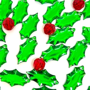 May include: A collection of green holly leaf-shaped ornaments with red jewel-toned accents. The ornaments are shiny and have a festive Christmas theme.
