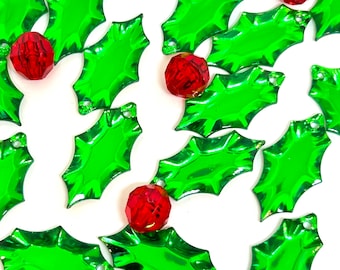 May include: A collection of green holly leaf-shaped ornaments with red jewel-toned accents. The ornaments are shiny and have a festive Christmas theme.