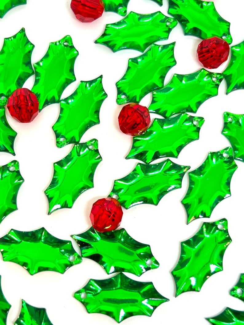 May include: A collection of green holly leaf-shaped ornaments with red jewel-toned accents. The ornaments are shiny and have a festive Christmas theme.