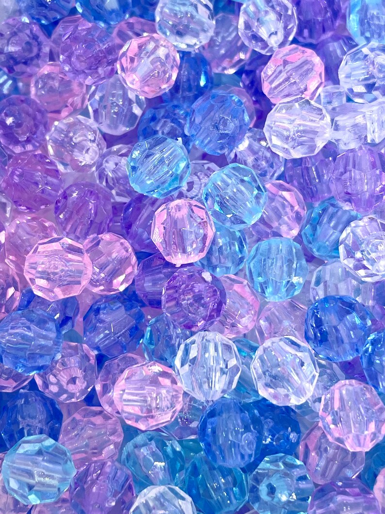 May include: A close-up of a pile of colorful faceted beads in shades of blue, purple, and pink. The beads are made of clear plastic and have a shiny finish.