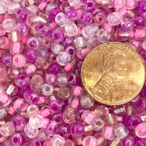K&ouml;nnte beinhalten: Ein Nahaufnahmebild eines gro&szlig;en Haufens kleiner, runder Glasperlen. Die Perlen sind eine Mischung aus klaren, schillernden, rosa und violetten Farben. Ein Cent-St&uuml;ck ist zur Gr&ouml;&szlig;enbestimmung enthalten.