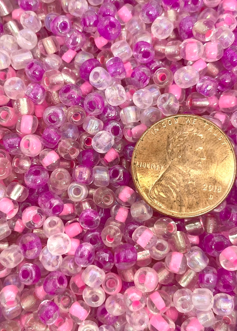 K&ouml;nnte beinhalten: Ein Nahaufnahmebild eines gro&szlig;en Haufens kleiner, runder Glasperlen. Die Perlen sind eine Mischung aus klaren, schillernden, rosa und violetten Farben. Ein Cent-St&uuml;ck ist zur Gr&ouml;&szlig;enbestimmung enthalten.