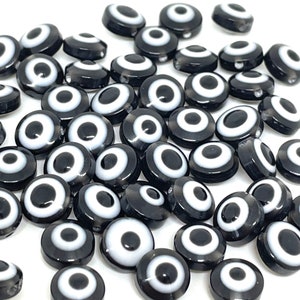 May include: A collection of black and white evil eye beads. Each bead is round with a black base, a white ring, and a black center, resembling an eye. The beads are scattered against a white background.