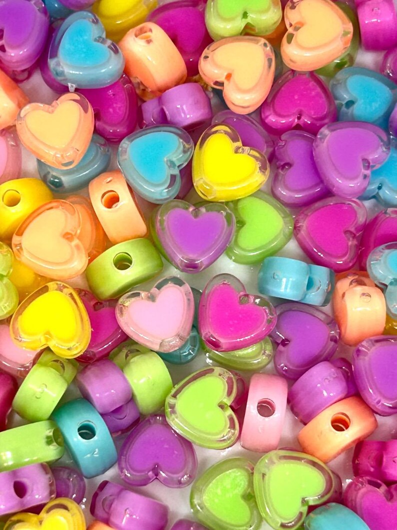 May include: A close-up of a variety of colorful heart-shaped beads. The beads are made of clear plastic with a solid color heart inside. The colors include pink, yellow, green, blue, and purple.