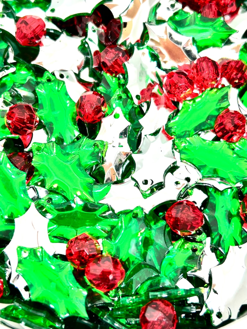 May include: A close-up of a pile of green and red plastic holly leaf charms. The charms are shiny and have a festive look.