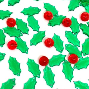 May include: Scattered green holly leaf-shaped beads and red round beads on a white background. The beads are made of a translucent material. Ideal for holiday crafts and decorations.