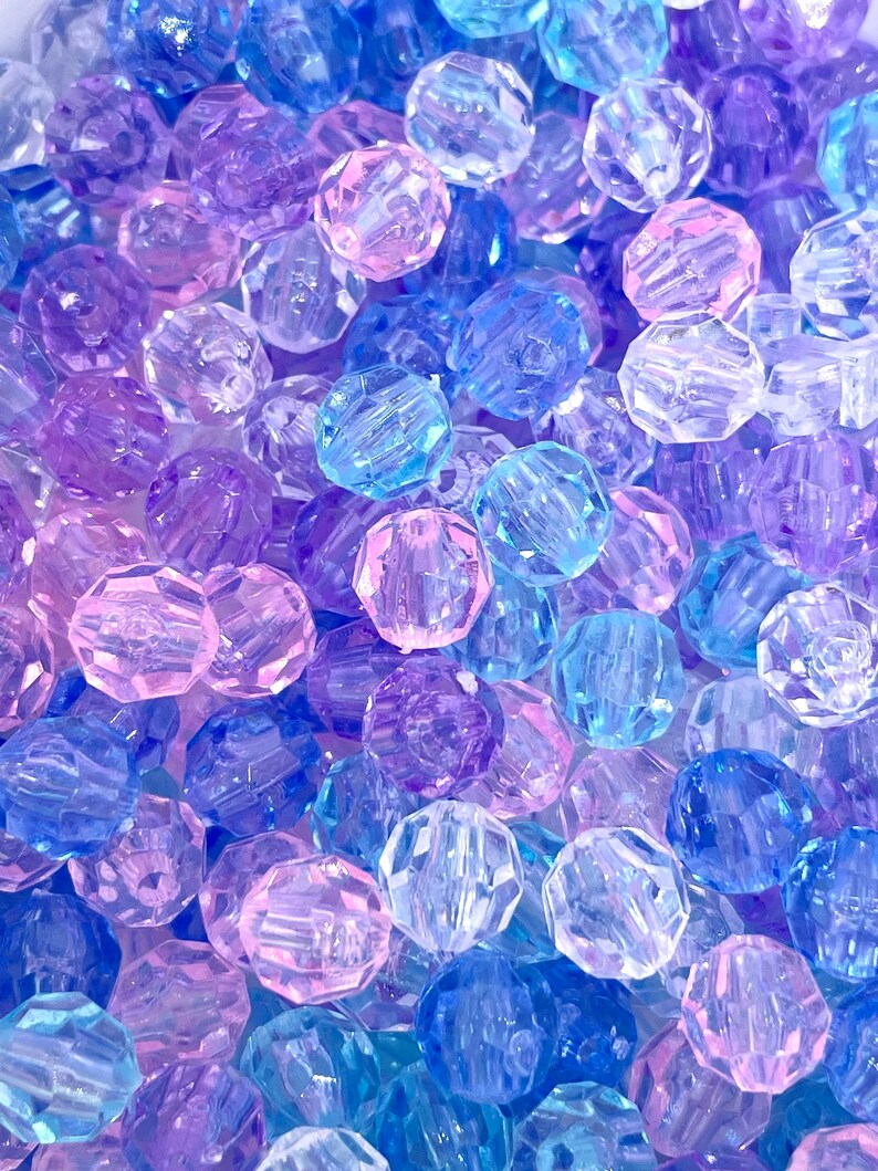 May include: A close-up of a pile of colorful, faceted, plastic beads. The beads are translucent and have a variety of shades of blue, purple, and pink.