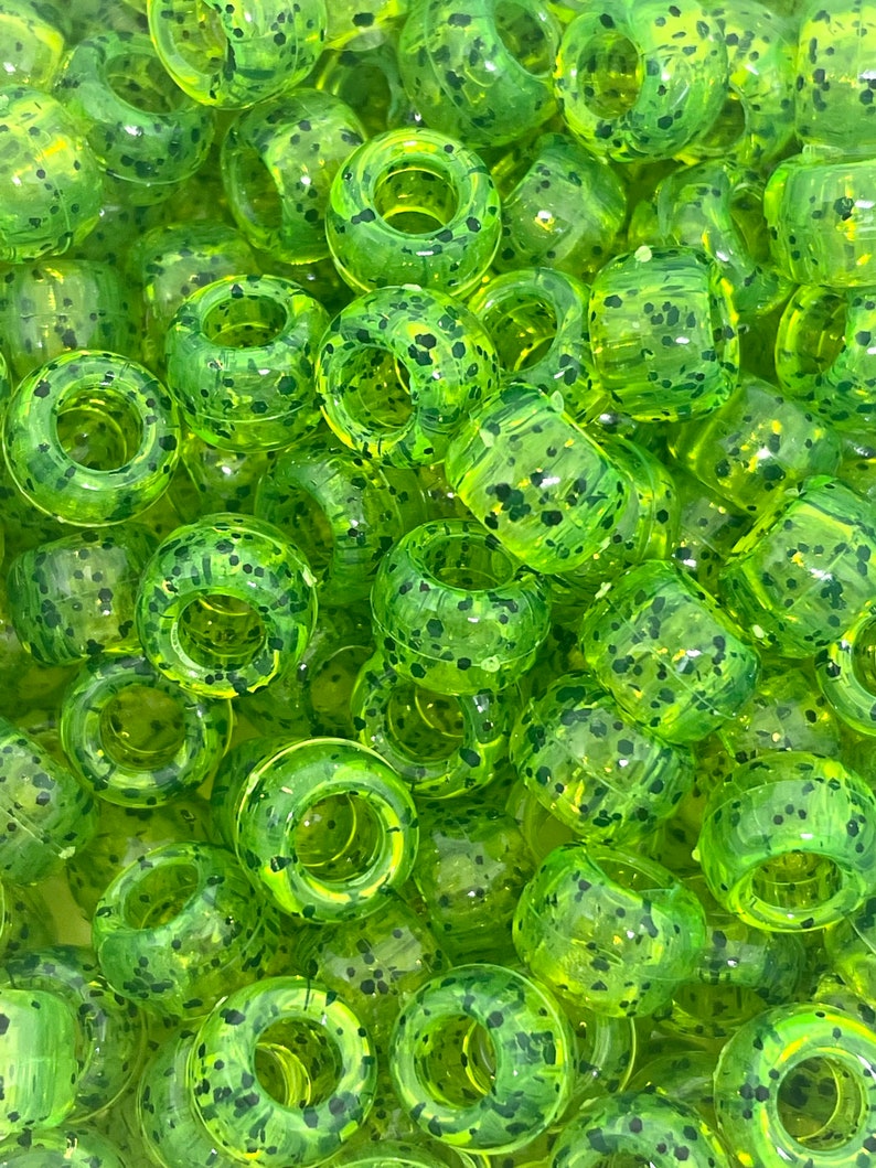 May include: A close-up of a large quantity of lime green and black speckled round beads with a hole in the center. The beads are made of plastic and have a glossy finish.