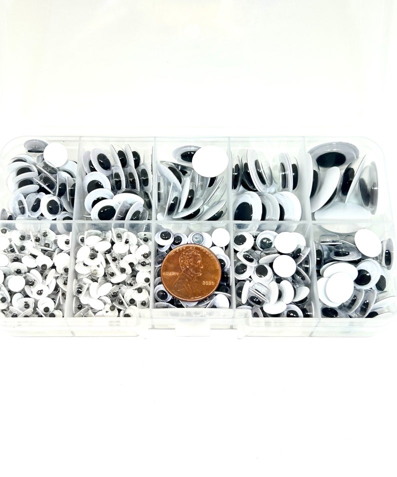 May include: A clear plastic storage box filled with various sizes of black and white googly eyes. The box has eight compartments and a penny is included for size reference.