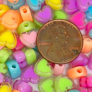 May include: A collection of colorful heart-shaped plastic beads with a hole in the center. The beads are translucent and have a shiny finish. The beads are scattered on a white surface. A US penny is included for scale.