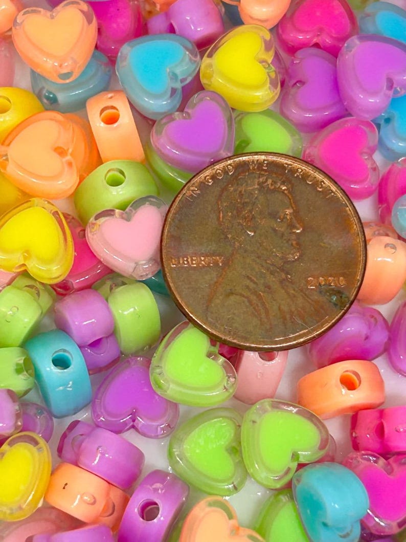 May include: A collection of colorful heart-shaped plastic beads with a hole in the center. The beads are translucent and have a shiny finish. The beads are scattered on a white surface. A US penny is included for scale.