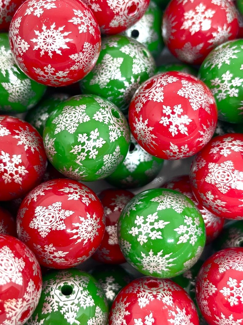May include: A close-up of a pile of Christmas ornaments. The ornaments are round and have a snowflake pattern on them. The ornaments are red and green. The snowflakes are silver.