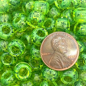 May include: A close-up of a pile of green and black speckled plastic beads. The beads are about the size of a US penny, which is shown in the image for scale.