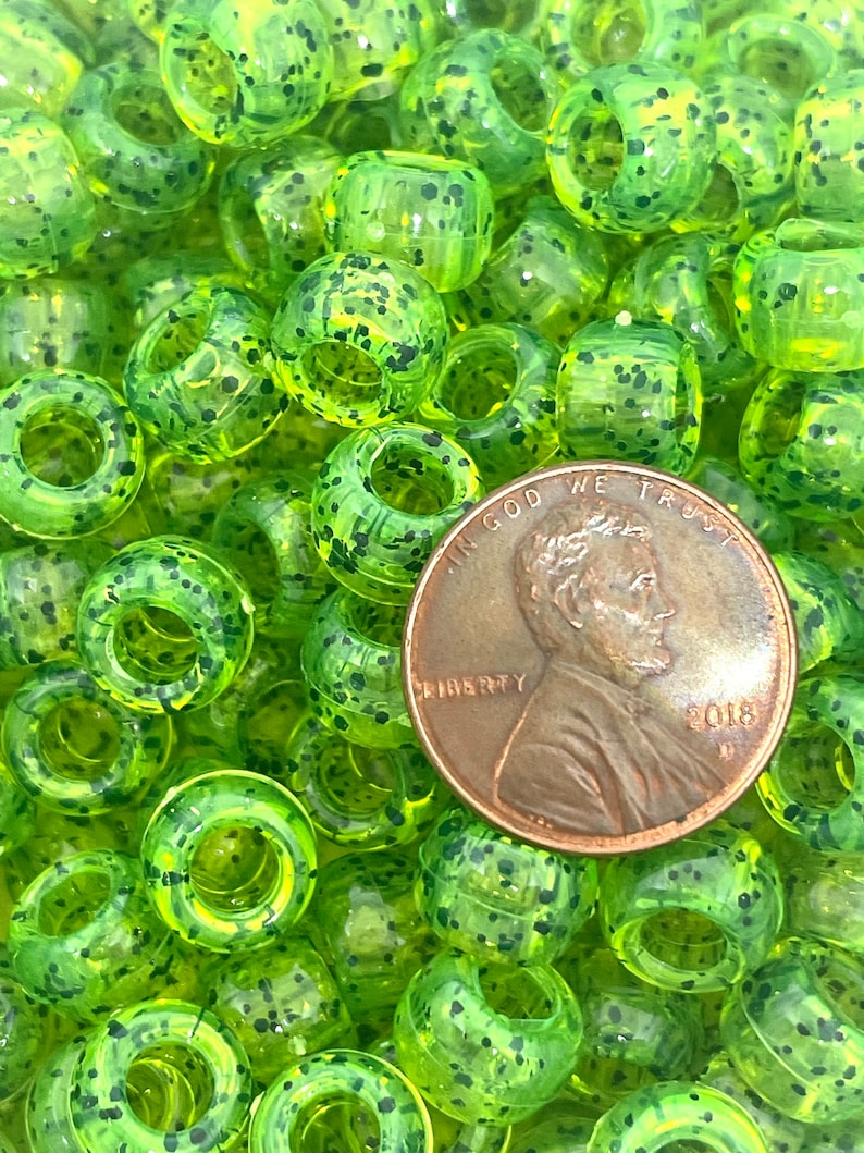 May include: A close-up of a pile of green and black speckled plastic beads. The beads are about the size of a US penny, which is shown in the image for scale.