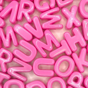 May include: A collection of glossy, bright pink plastic alphabet letters scattered in a random arrangement. Each letter features a smooth, rounded, three-dimensional shape with a shiny finish, creating a dense pattern of various characters.