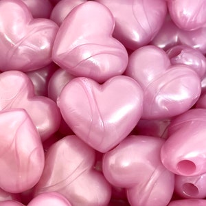 May include: A close-up of a pile of pink heart-shaped beads. The beads have a pearlescent finish and are made of plastic.