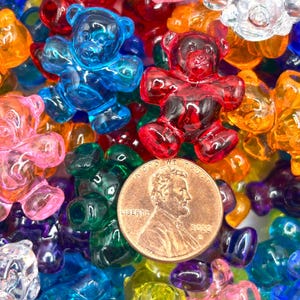 May include: A close-up of a collection of colorful, translucent gummy bear-shaped beads. The beads are in various colors, including blue, red, pink, green, yellow, and orange. A US penny is in the center of the image.