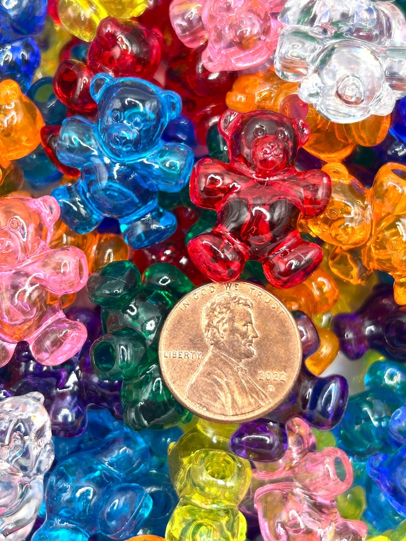May include: A close-up of a collection of colorful, translucent gummy bear-shaped beads. The beads are in various colors, including blue, red, pink, green, yellow, and orange. A US penny is in the center of the image.