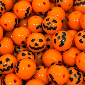 K&ouml;nnte beinhalten: Nahaufnahme von orangefarbenen K&uuml;rbis-Themen-Perlen mit schwarzen Gesichtern. Diese runden Perlen haben eine gl&auml;nzende Oberfl&auml;che und sind mit verschiedenen Jack-o'-Lantern-Gesichtern versehen. Perfekt f&uuml;r Halloween-Bastelarbeiten und Schmuckherstellung.