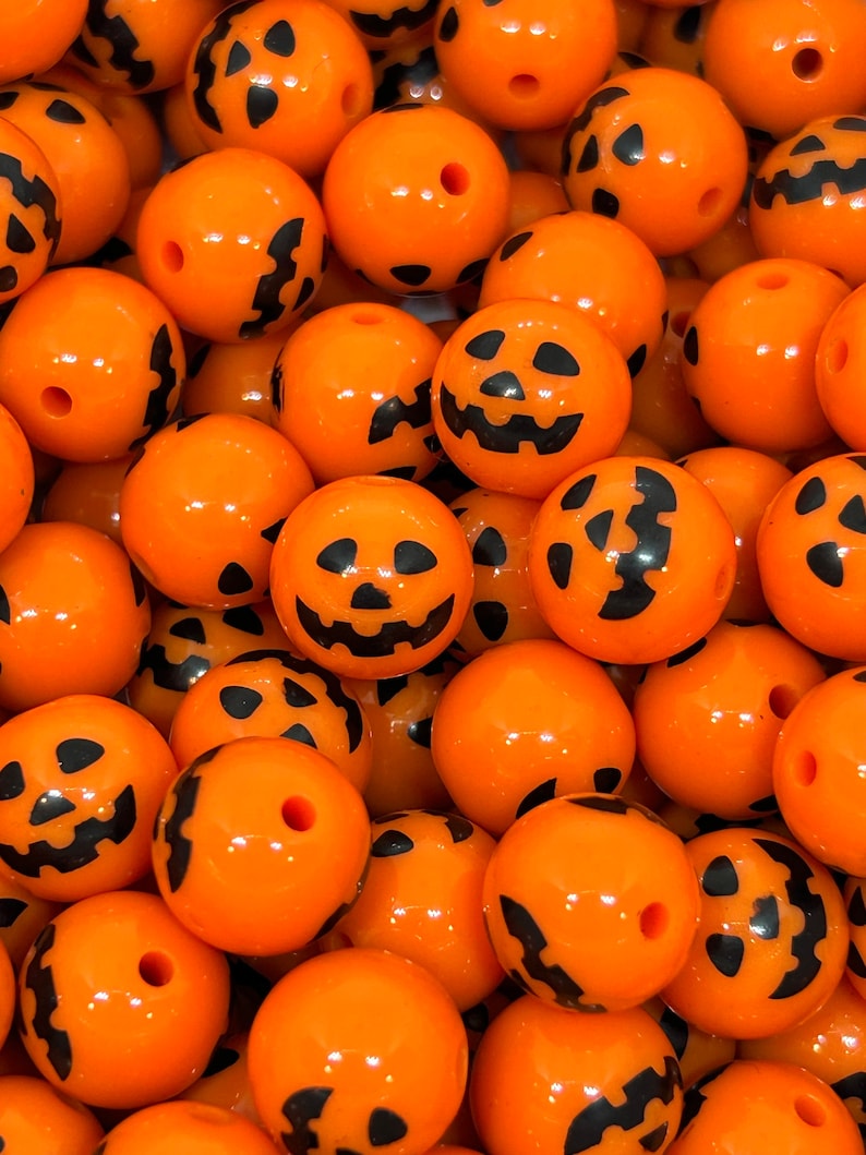 K&ouml;nnte beinhalten: Nahaufnahme von orangefarbenen K&uuml;rbis-Themen-Perlen mit schwarzen Gesichtern. Diese runden Perlen haben eine gl&auml;nzende Oberfl&auml;che und sind mit verschiedenen Jack-o'-Lantern-Gesichtern versehen. Perfekt f&uuml;r Halloween-Bastelarbeiten und Schmuckherstellung.