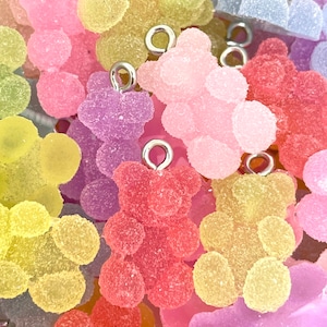 May include: A close-up of a group of colorful gummy bear charms. The charms are made of a translucent, textured material and have a shiny finish. Each charm features a small silver loop for attaching to a necklace, bracelet, or other jewelry.