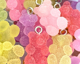 May include: A close-up of a group of colorful gummy bear charms. The charms are made of a translucent, textured material and have a shiny finish. Each charm features a small silver loop for attaching to a necklace, bracelet, or other jewelry.