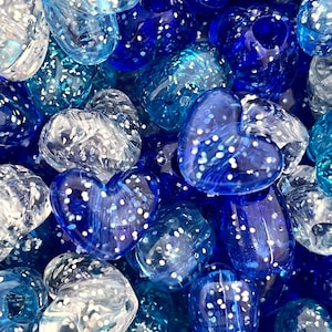 May include: A close-up of a pile of blue and white heart-shaped glass beads. The beads are covered in white glitter and have a translucent appearance.