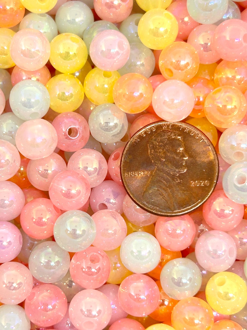 May include: A close-up image of a large assortment of iridescent pastel-colored beads. The beads are round and have a smooth, glossy finish. The beads are arranged in a random pattern, creating a visually appealing display.