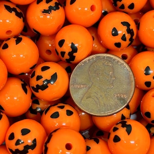 K&ouml;nnte beinhalten: Nahaufnahme von orangefarbenen Perlen mit schwarzen K&uuml;rbisgesichtern. In der Mitte befindet sich eine M&uuml;nze mit dem Jahr 2020. Die Perlen sind rund mit einem Loch in der Mitte, ideal f&uuml;r Schmuck oder Halloween-Bastelarbeiten.