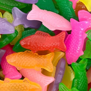 May include: A close-up shot of a pile of colorful, translucent gummy fish candies. The fish are in various colors, including pink, orange, red, green, purple, and yellow. The candies have a glossy appearance and are scattered randomly.