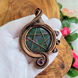 May include: A copper wire wrapped pendant featuring a green and pink star-shaped stone with a five-pointed star design. The pendant is accented with four small purple stones.