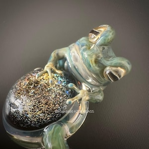 Puede incluir: Una escultura de rana de cristal en tonos azules, verdes y dorados, posada sobre un orbe moteado. Los ojos y las patas de la rana tienen detalles dorados. Una pieza de arte única.