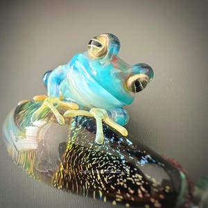 Puede incluir: Escultura artesanal de rana de vidrio en tonos azules, amarillos y transparentes. La rana está posada sobre una base de vidrio transparente con un patrón iridiscente moteado. Diseño artístico y caprichoso.
