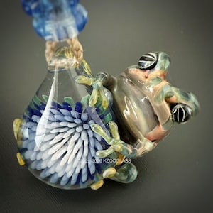 Peut inclure: Un pendentif en verre représentant une grenouille sculptée à la peau verte et brune translucide, agrippée à un récipient en verre transparent. Le récipient contient un motif floral bleu et blanc. Le texte 2021-2026 KZOOGLASS est visible.