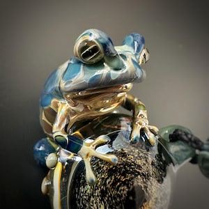 Puede incluir: Una detallada escultura de rana de vidrio en tonos azules, dorados y verdes. La rana está posada sobre una base texturizada, mostrando detalles intrincados y un acabado brillante. La escultura tiene el texto "©2021-2020 ZING GLASS".